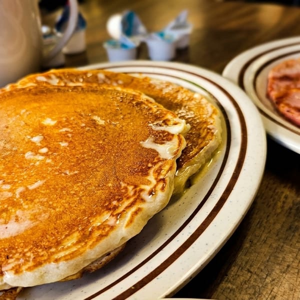 Golden pancakes with syrup and butter, alongside ham and eggs on a wooden table.