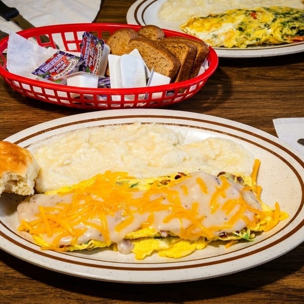 Two plates of breakfast food, including omelets, grits, biscuits, and toast, on a wooden table.