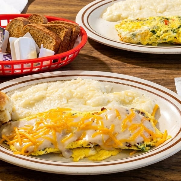 Two plates of omelets with grits, toast, and a biscuit on a wooden table.