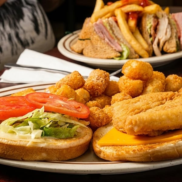 A fried fish sandwich with tater tots and a club sandwich with fries on a wooden table.