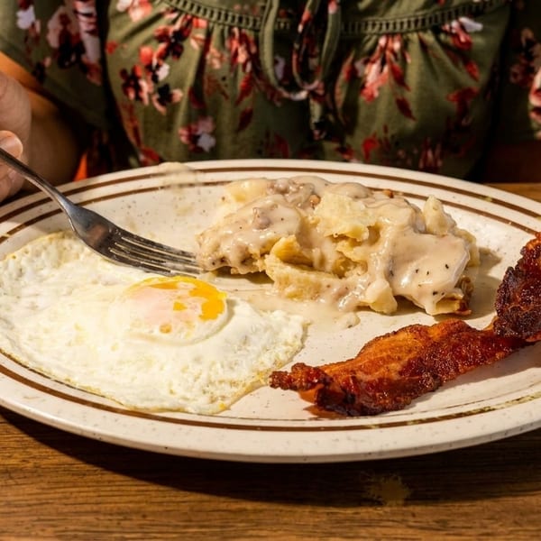 A person eating a breakfast plate with fried eggs, bacon, and biscuits with gravy.